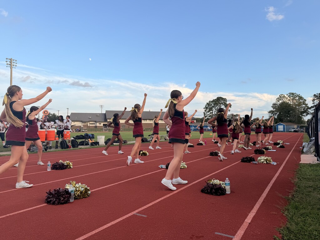 Group of cheerleaders practicing on a red track, some holding pom-poms, others cheering. Background includes grass and distant trees.