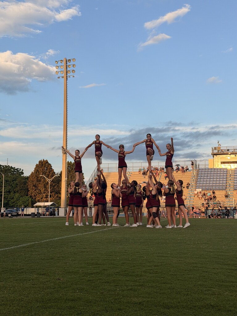 Cheerleaders in maroon uniforms perform stunts on a field with a stadium in the background.