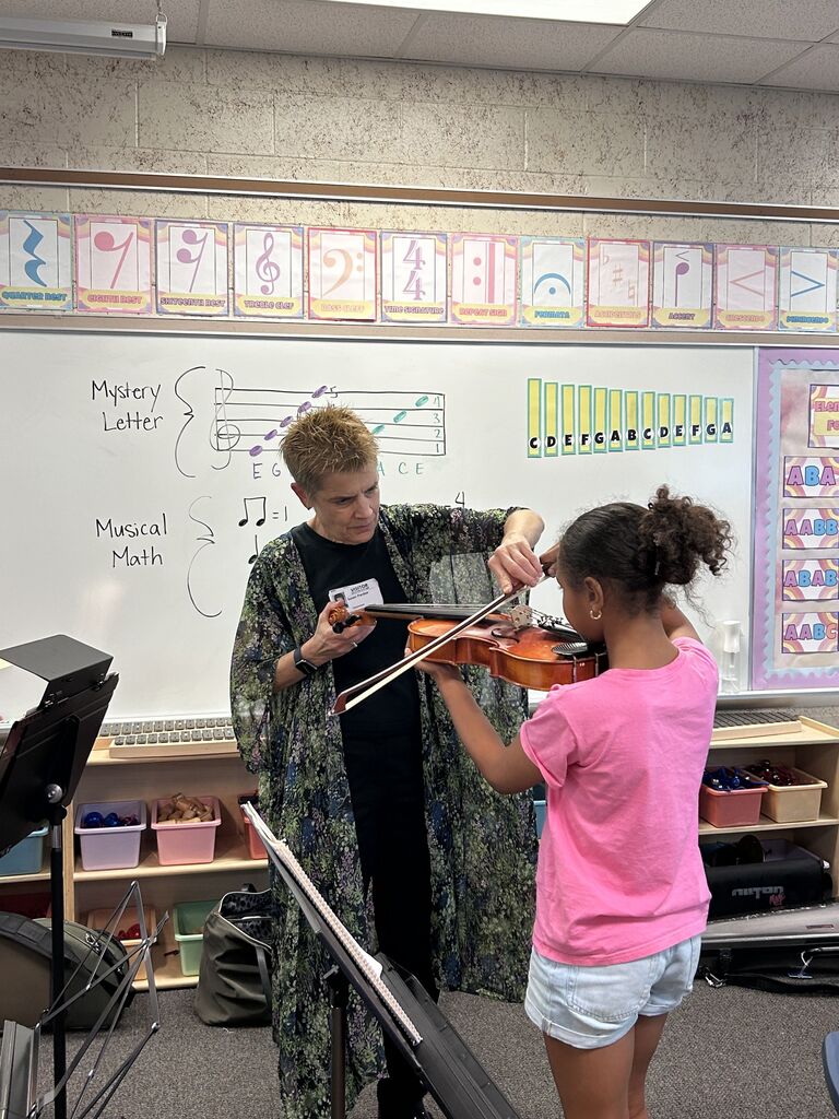 Teacher helping young student play violin. Bright white classroom with whiteboard and shelves in background.