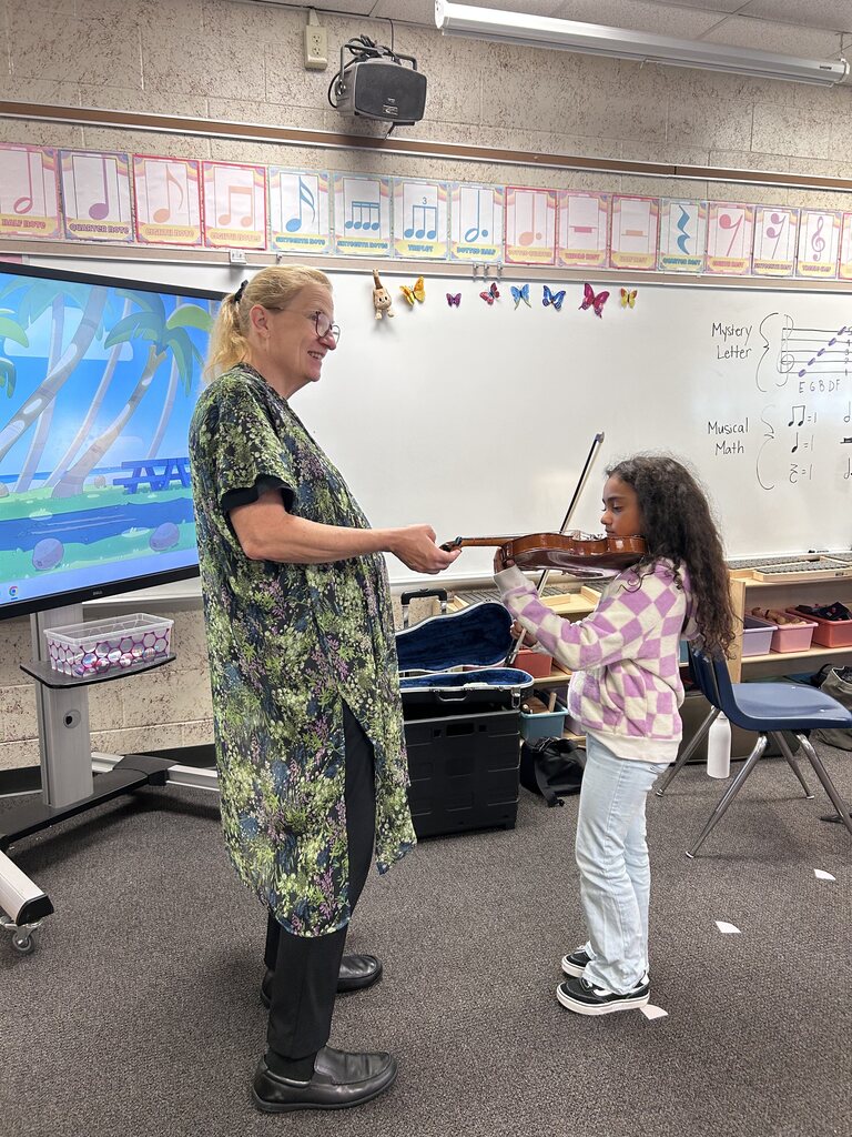 Teacher showing girl how to play violin in classroom. Teacher holding violin bow.