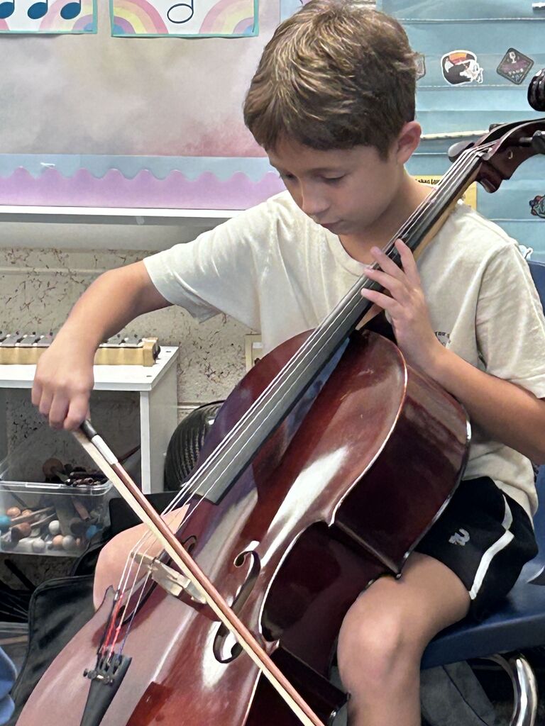 A boy with blond hair plays a cello while seated in a chair. He holds a bow in his right hand.