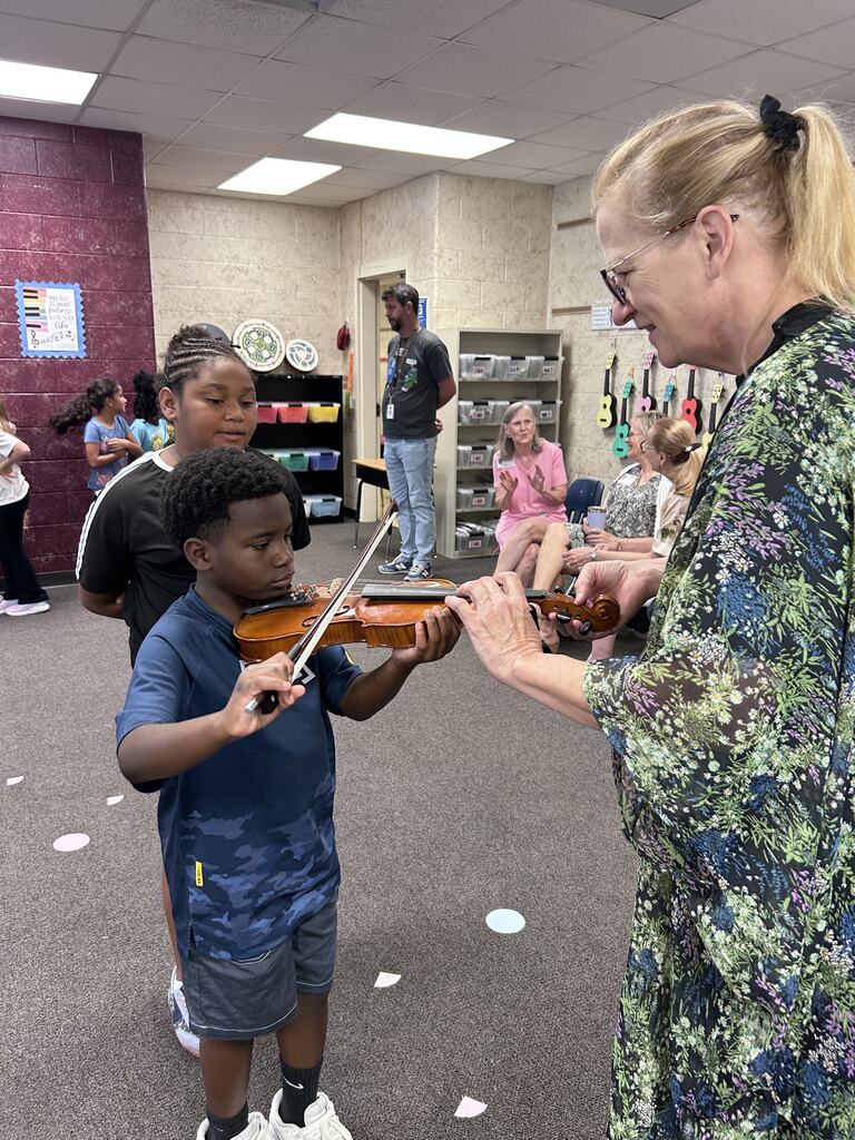 Woman helping a boy play violin in a classroom. A few students stand behind them.