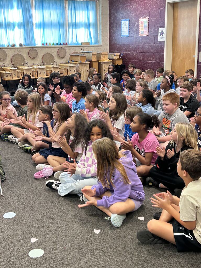 A group of children sits on the floor, clapping, in a classroom with blue blinds and a purple wall.