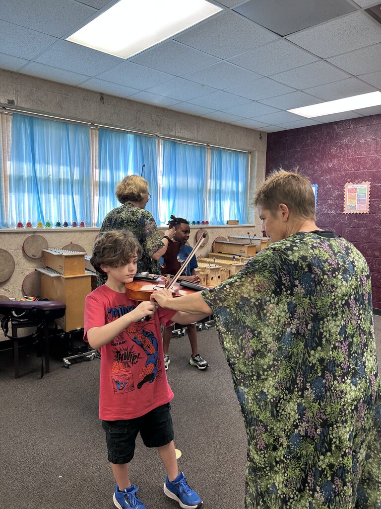 A teacher guides a boy playing violin in a room with blue curtains, seated individuals, and instruments.