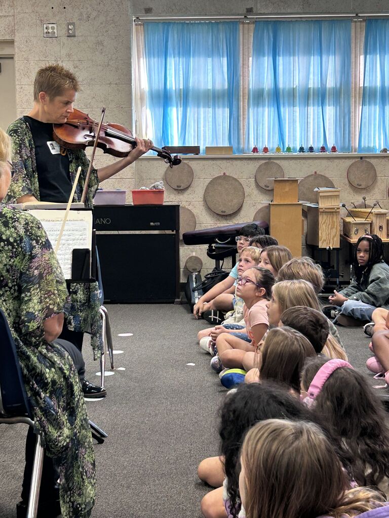 A violinist plays for a seated audience in a classroom with blue drapes, a piano, and a drum set in the background.