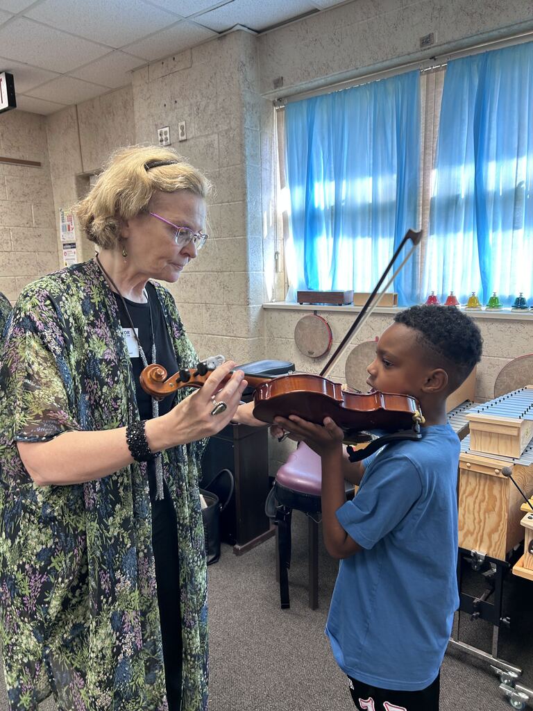 A woman with glasses shows a young boy a violin in a room with a blue backdrop.