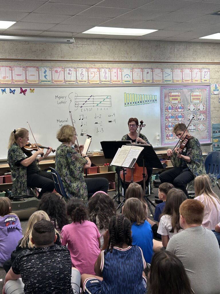 Four violinists perform in a classroom. Audience seated on the floor. Whiteboard with various colorful educational posters in background.