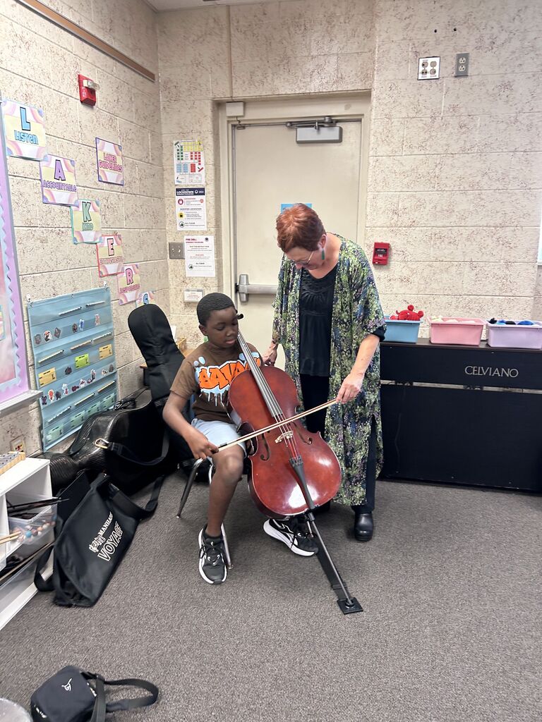 A seated student plays a cello, with a teacher assisting. The room has a beige wall, a door, and various classroom items.