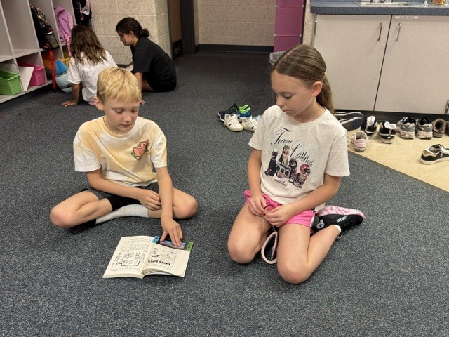 Two children are sitting on a carpet, one reading a book, while others sit around them.