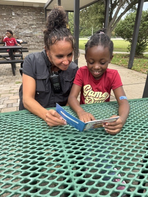 An adult and child sit at a table reading a blue book. Behind them is a brick wall, picnic tables, and trees.