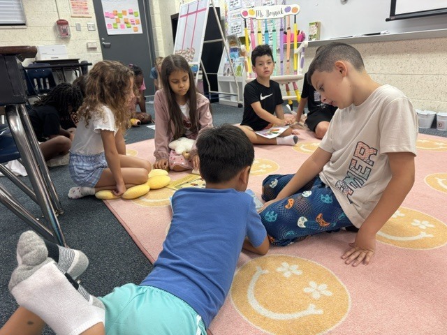 Children sit on a carpeted floor, reading books. Some have stuffed toys nearby. A boy in a white shirt leans over another boy.