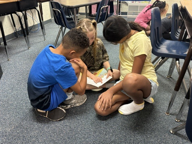 Four children are gathered on a classroom floor, with two reading from a book. The room has chairs and tables.