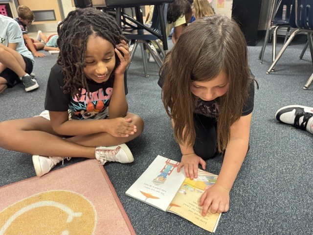 Two children on a floor with a rug, reading a book together. Other children are seated in the background.