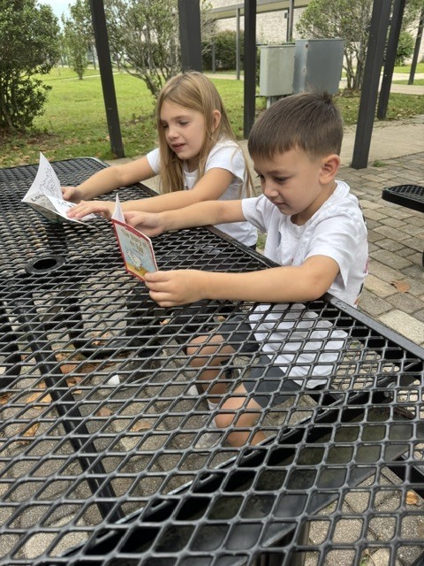 Two children seated at a table, reading a book. One child holds a book, and the other holds a paper.