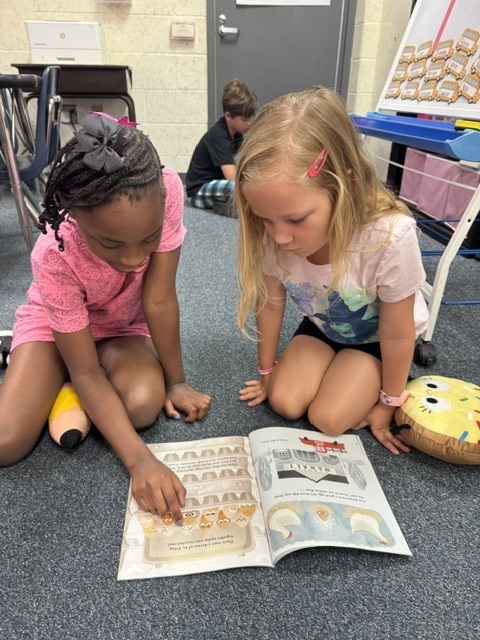 Two young girls sitting on the floor, one holding a pencil, reading a book together.