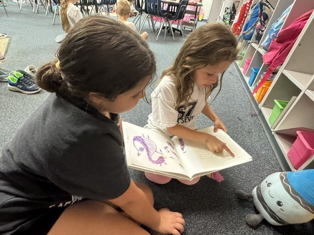 Two children, one holding a book, read together. One wears a gray t-shirt, and the other wears white.