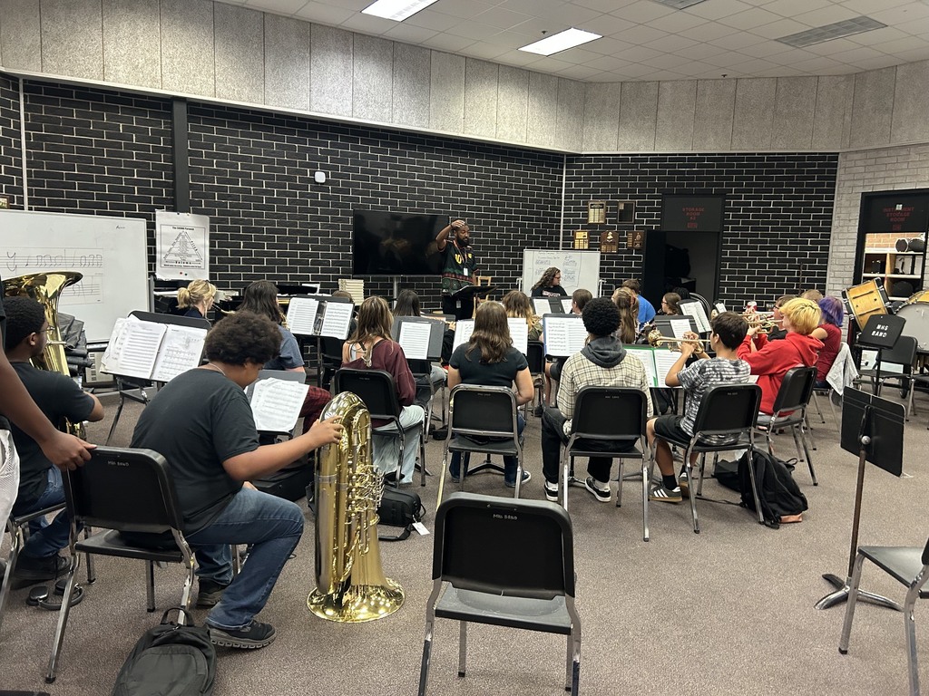 A band class in a room with black walls. Students sit in chairs with instruments. A teacher stands near a screen.