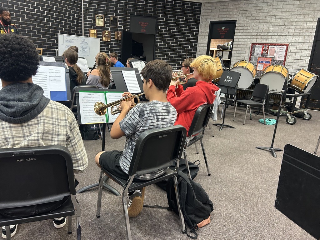 People in a classroom are sitting and playing instruments. One person is playing a trumpet.