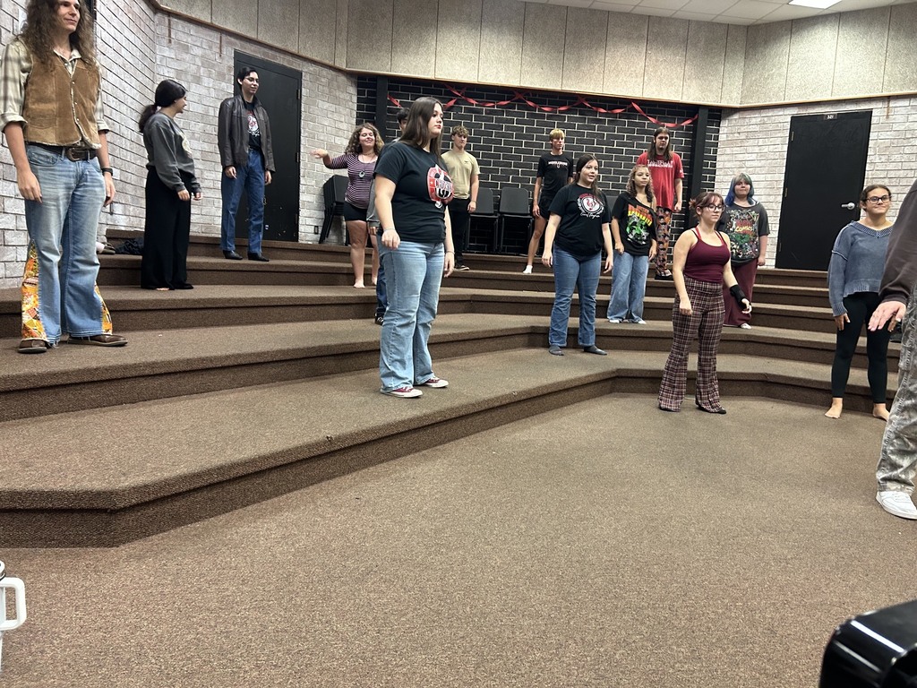 A group of people dressed casually stand on various levels of a stage. Brick walls and a high ceiling are visible.