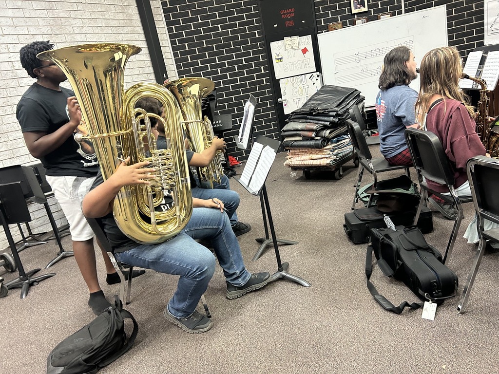 A group of people sit in chairs, playing large brass instruments. Two individuals play tuba, while others play trombone.
