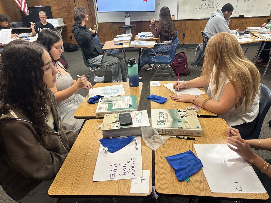 Students seated at desks in a classroom, engaged in a writing activity. Books and papers on the desks, and a whiteboard in the background.