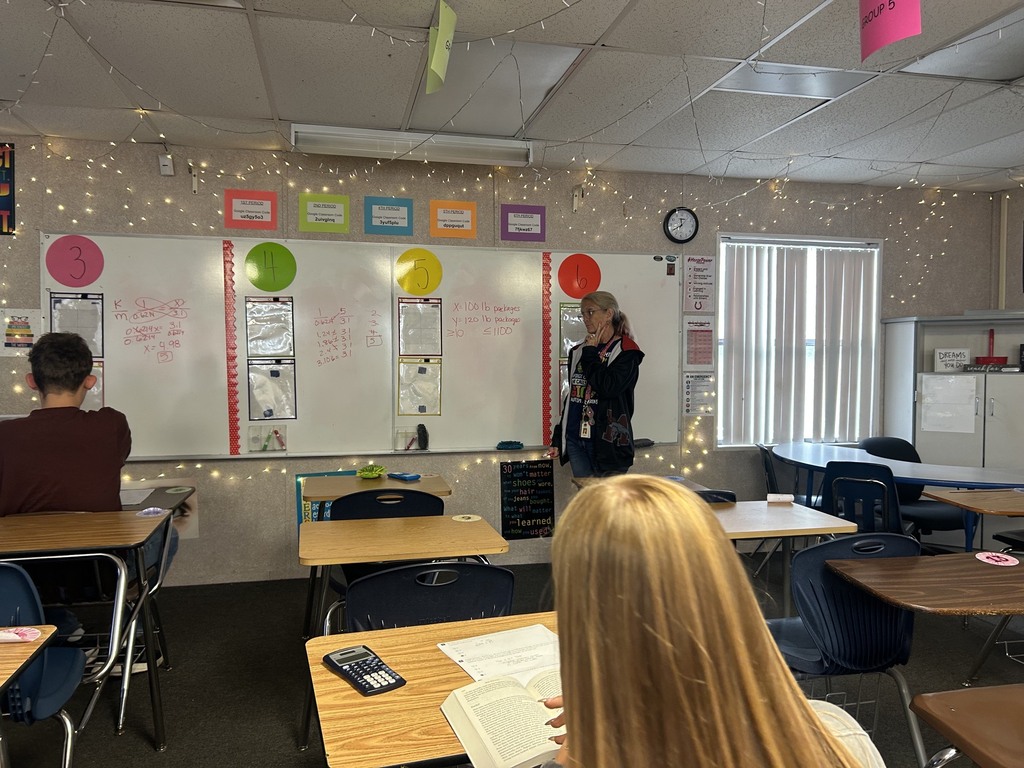 A classroom with desks, chairs, a teacher, and students. The walls have a whiteboard, decorations, and a clock.