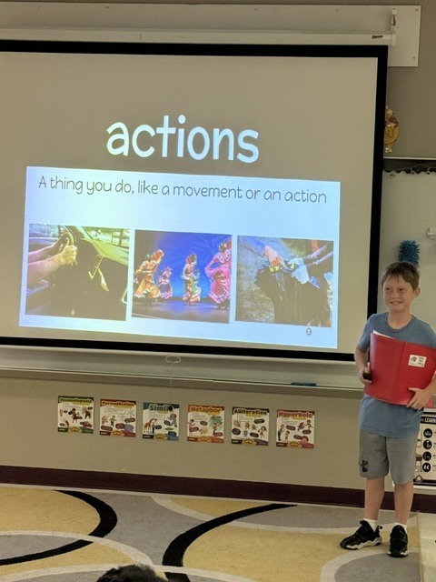 Classroom with a projector screen displaying "actions" and a boy standing, holding a red folder.