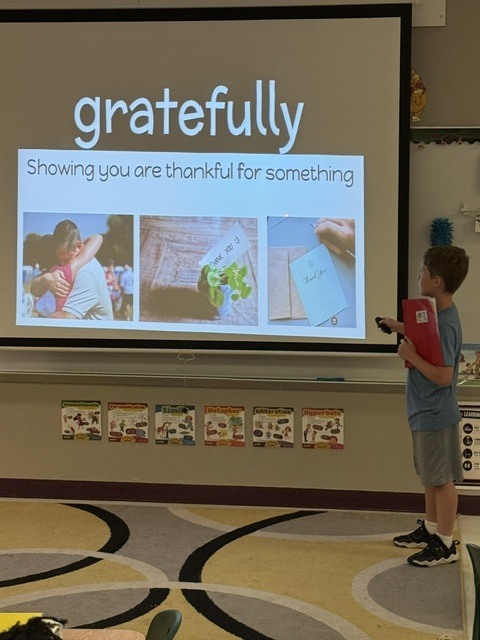 Child stands by a screen with 'gratefully' and three images, holding a red folder, in a classroom.