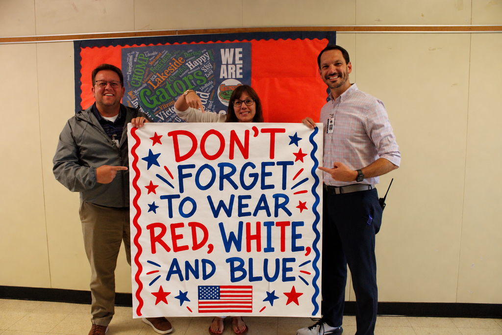 Three people hold a sign with "Don't Forget to Wear Red, White, and Blue" in front of a wall.