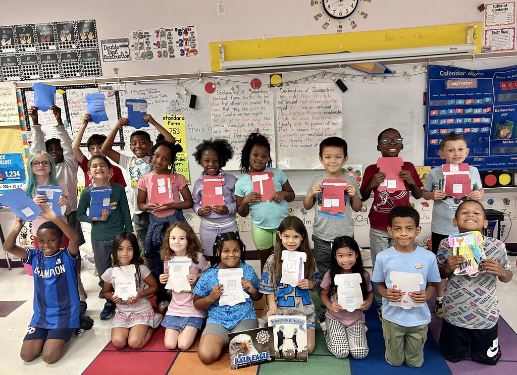 Classroom with a group of students, some standing and others sitting, holding up certificates and books.