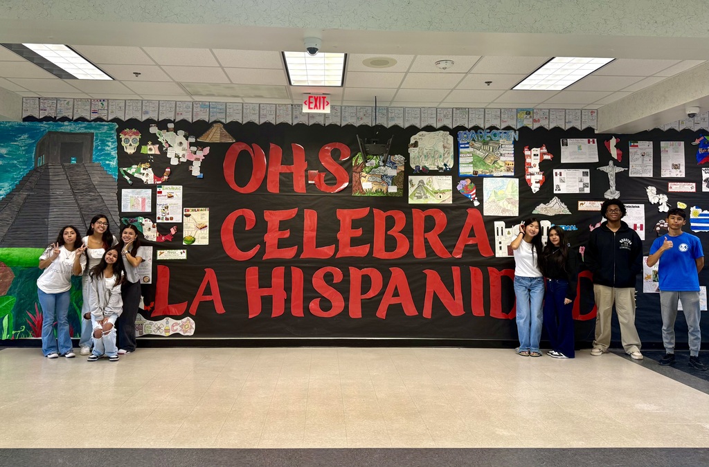 A group of students standing together in front of a large poster that says "OHS CELEBRA LA HISPANIDAD".