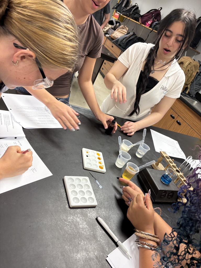 Three women in lab coats conduct an experiment at a lab table. Papers, a pipette, and small containers are on the table.