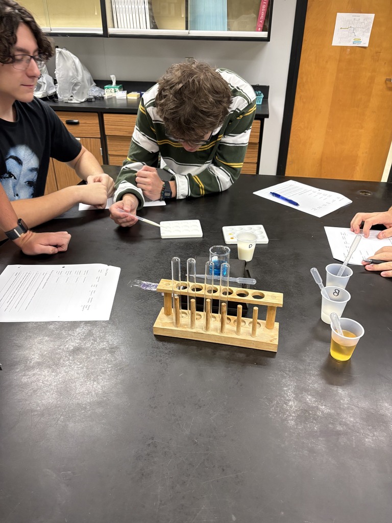 Students gather around a table with scientific equipment, papers, and a yellow cup. One student holds a pipette.