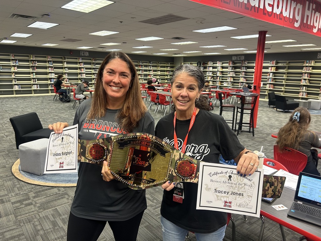 Two women in library, holding certificates and championship belt, smiling. Background includes chairs, desks, and bookshelves.