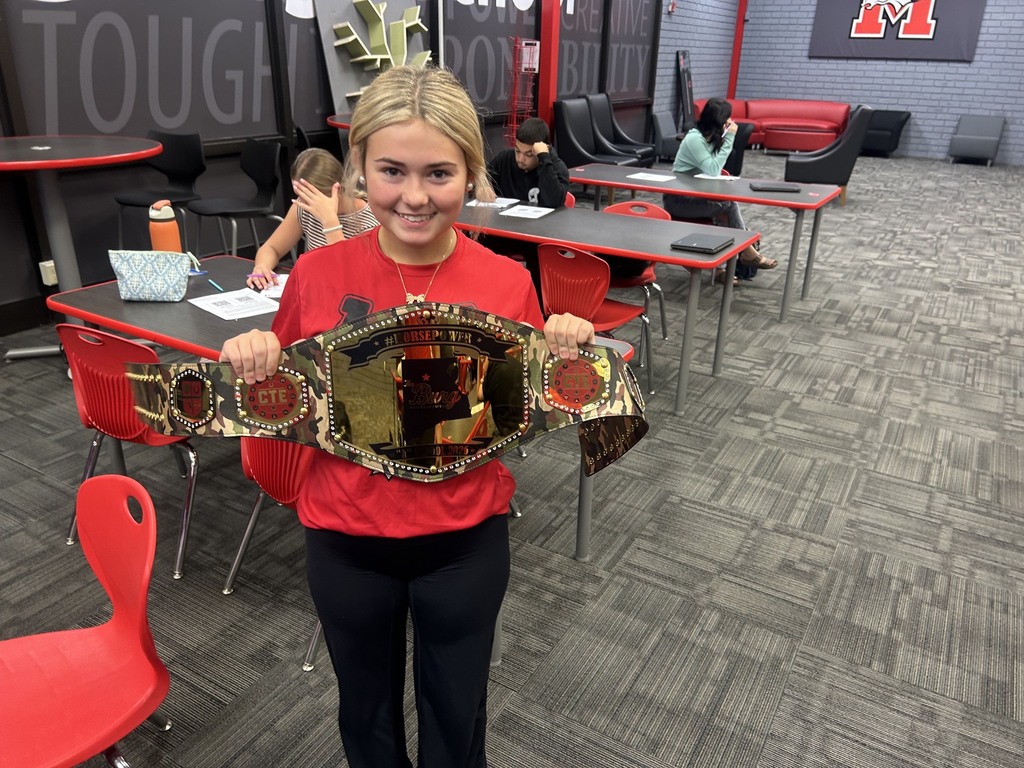 Woman with a belt in a classroom. Background includes tables, chairs, and a red wall with the word "Tough".