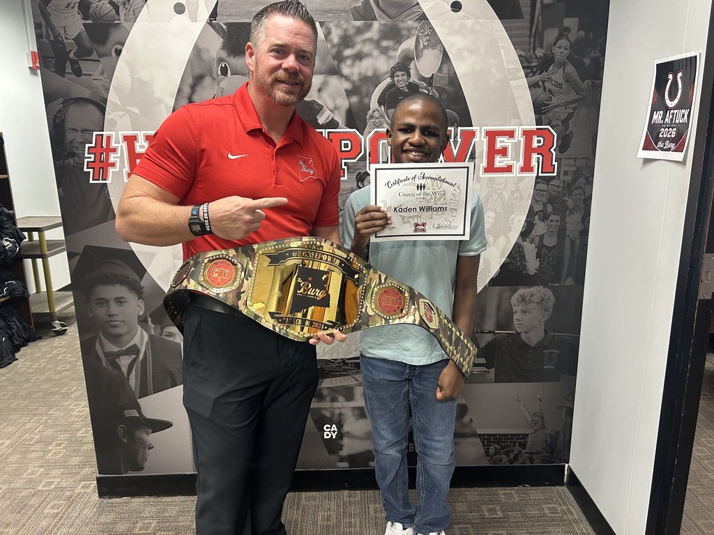 Two men stand in a room with a sports-themed wall in the background. One holds a championship belt and points at it.