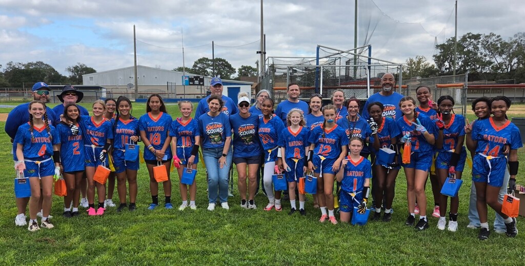A group of people in blue uniforms stands on a grassy field, holding orange bags. A fence and trees are in the background.