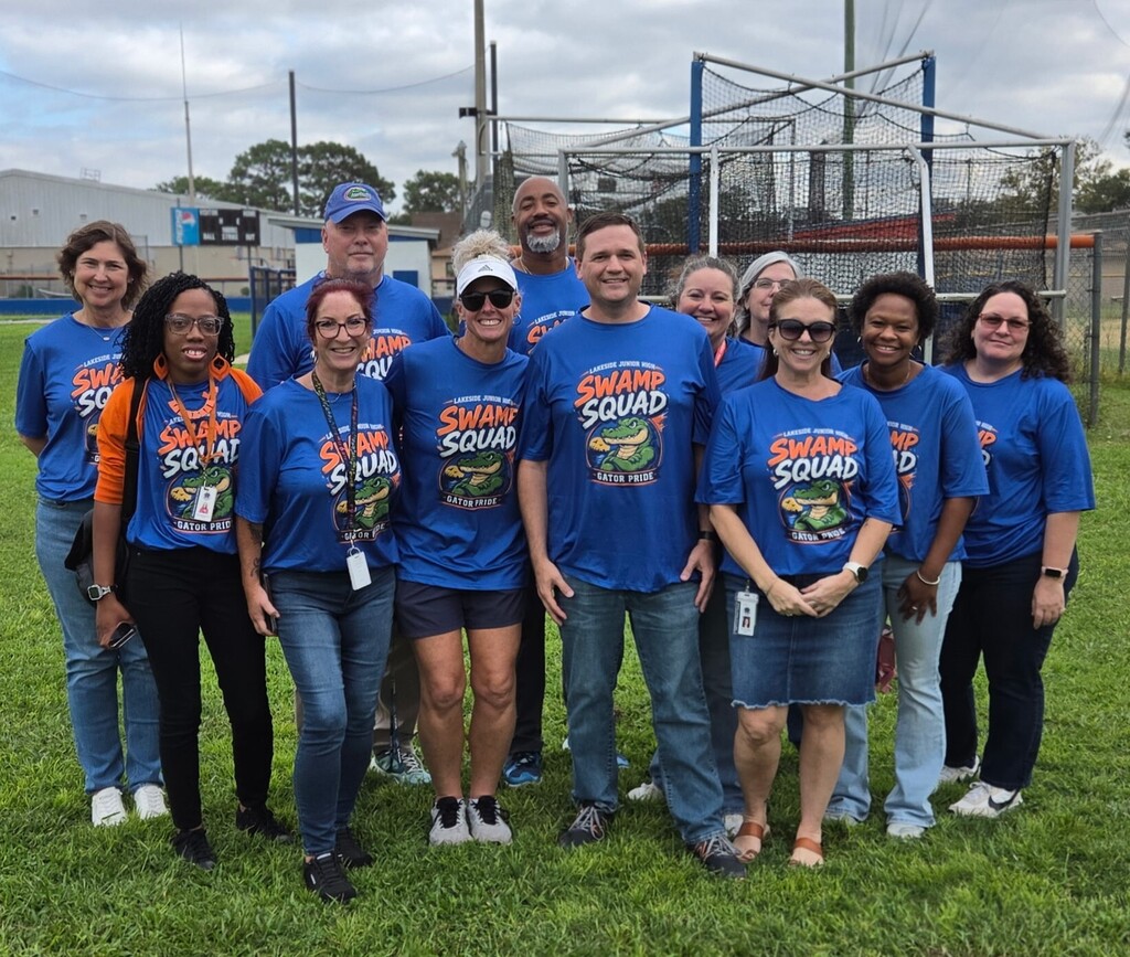 Group of adults in blue shirts, some wearing hats, pose for a photo on a grassy field with a batting cage in the background.