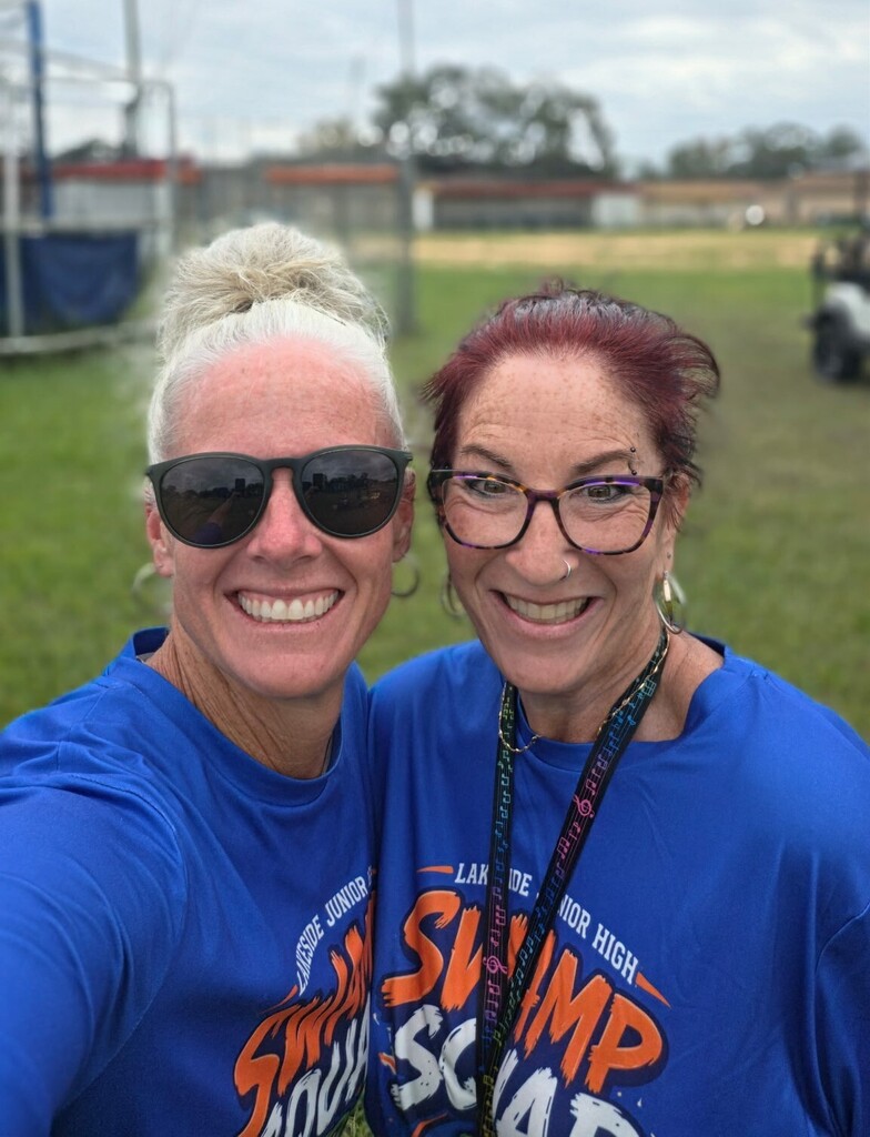Two women in blue shirts with "Lafayette Swamp" and "High School" print stand on a field, smiling for a selfie.