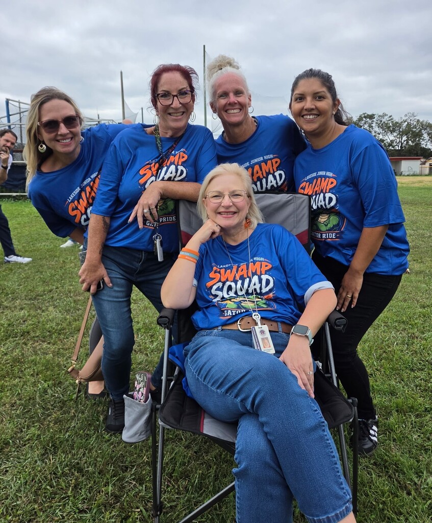 Five women in matching blue t-shirts stand and sit on a grassy field. The woman in the middle wears jeans and a watch.