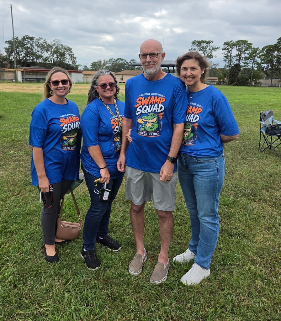 Four people wear blue Swamp Squad shirts and stand on a grassy field with trees in the background.