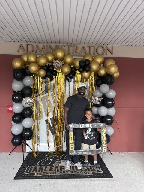 A man and child stand in front of a balloon arch. The arch has balloons and gold streamers.