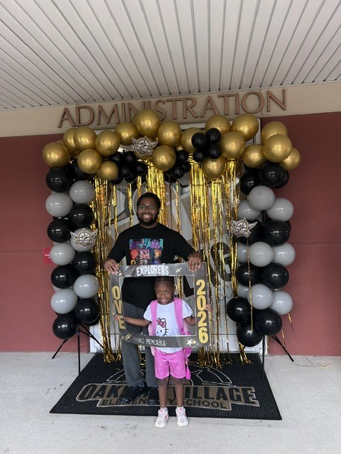A man and a young girl stand in front of a backdrop with "2026" and balloons. They are at a school.