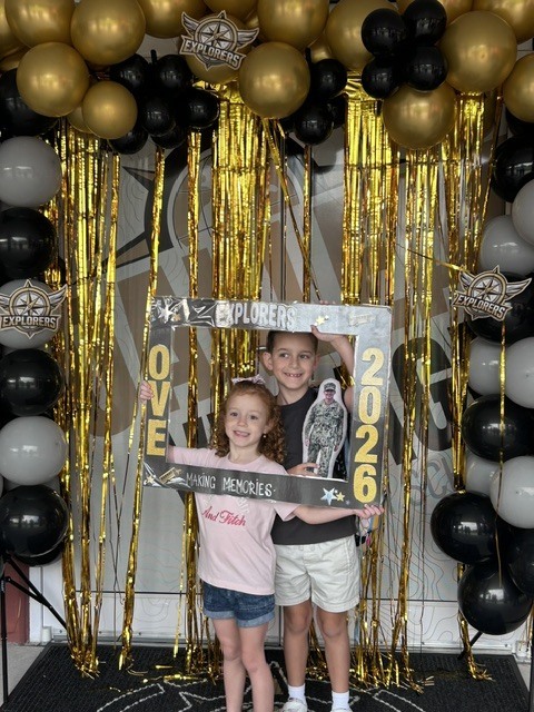 Two children stand in front of a black-and-gold balloon arch and a sign that reads "LOVE 2026."
