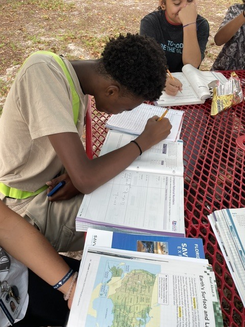 Two students in outdoor area, one writing on a notebook, other reading. Table with books, papers, and a map.