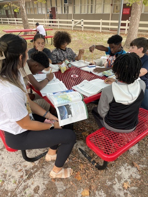 A group of children and an adult sit at a picnic table with books and papers.