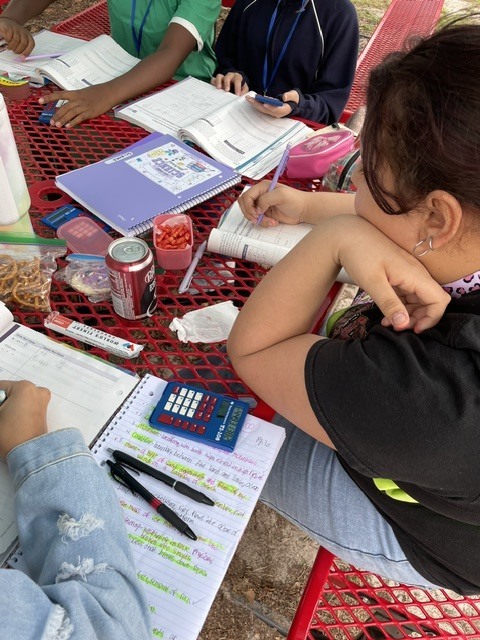 Students seated around a table, using notebooks and pens. Various items like calculators, books, and snacks are present.