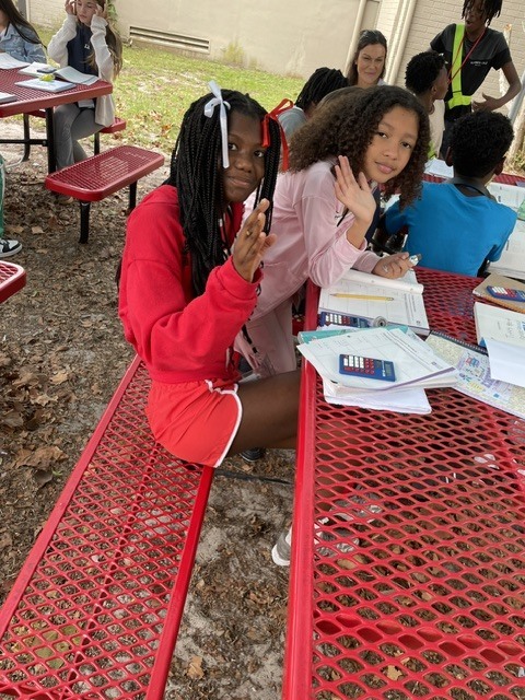 Two students in red clothes sit at a picnic table, holding books and papers. Nearby, others are seated.