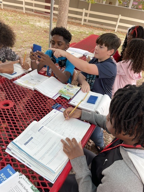 Students sitting at a red picnic table outdoors, some using cell phones, others writing in books, with a red fence and tree in the background.