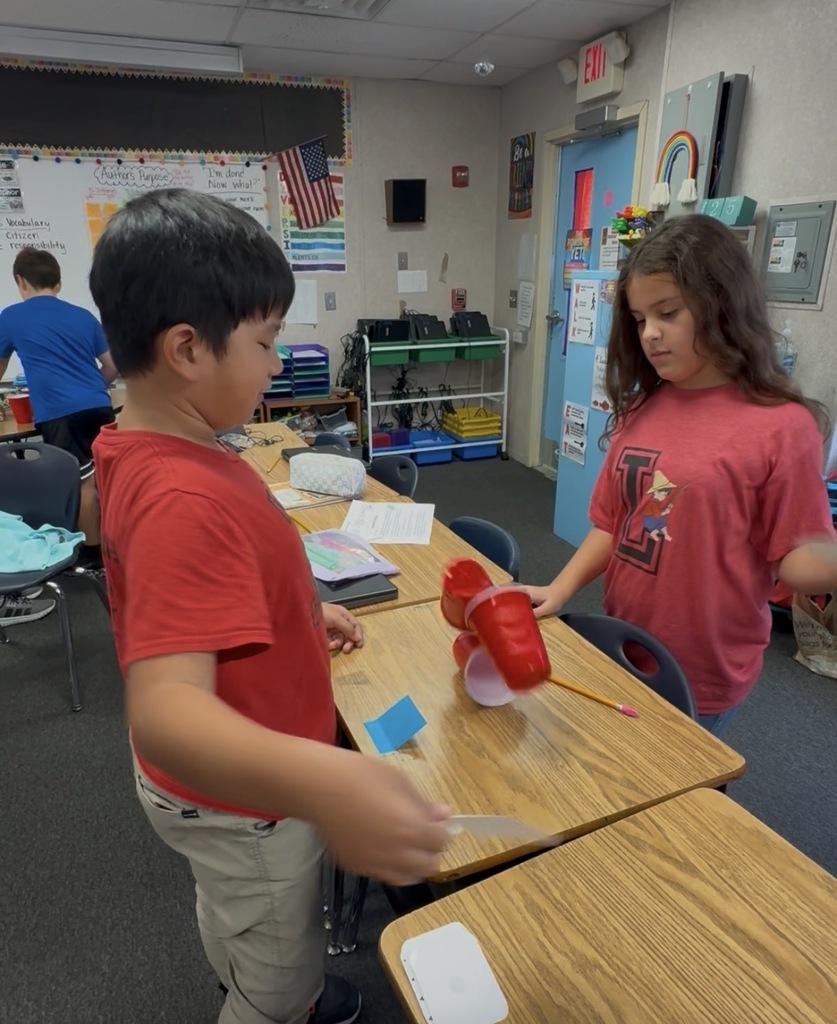 Children at a table, one holding paper and scissors, another holding a red cup.